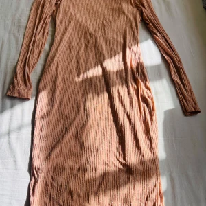 Glittrig långklänning i persikoton - Säljer en långklänning i persikofärgad ton med glittriga ränder och v-ringning. Klänningen har lång ärm och är tillverkad i ett stretchigt, glittrigt material som ger en snygg effekt. Perfekt för dig som vill sticka ut med stil.