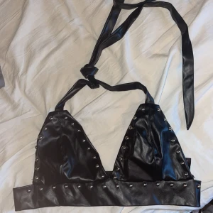 Svart faux läder korsett-topp med nitar från Romwe - Säljer en svart korsett-topp i faux läder från Romwe. Toppen har halterneck-band att knyta i nacken och är dekorerad med silvriga nitar längs kanterna. Perfekt för en edgy look!