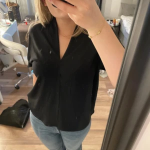 Svart blus med v-ringning - En stilren svart blus med v-ringning och korta ärmar. Passformen är normal och tyget känns mjukt och följsamt. Perfekt att matcha med jeans eller kjol för en enkel men snygg look.