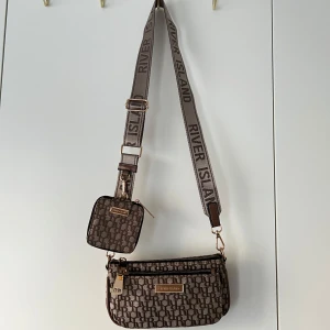 Brun axelremsväska från River Island - Säljer en brun axelväska från River Island med matchande liten plånbok på remet. Väskan har justerbar axelrem med logga, två dragkedjor och mönster med RI-bokstäver över hela ytan. Perfekt för att bära det viktigaste med stil. Priset kan diskuteras. 