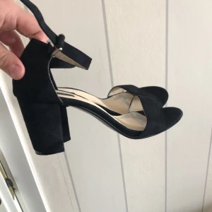 Svarta klackskor med ankelrem - Säljer ett par svarta klackskor med öppen tå och bred klack. Skorna har en stilren design med rem över tårna och justerbar ankelrem. Perfekta för dig som vill ha både stil och komfort.