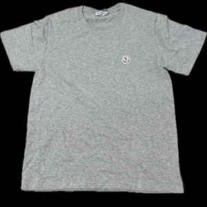 Grå t-shirt från Moncler - Säljer en stilren grå t-shirt från Moncler med liten broderad logga på bröstet. T-shirten har klassisk passform och är tillverkad i mjuk bomull. Perfekt för en enkel och clean look.