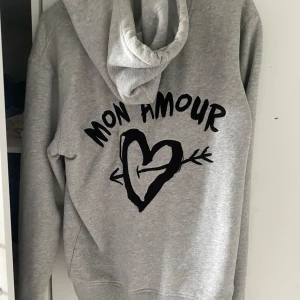 Grå hoodie med tryck från Mira Paris - Säljer en grå hoodie från Mira Paris med svart tryck på ryggen där det står 'Mon Amour' och ett hjärta med pil. Tröjan har huva med dragsko och en stor magficka framtill. Perfekt för en avslappnad stil. Knappt använd 
