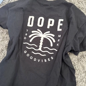 Svart t-shirt från Dope med palmtryck - Svart t-shirt från Dope med vit print på både bröstet och ryggen. Trycket har en palm, vågor och texten 'GOODVIBES'. Klassisk passform och rund halsringning. Perfekt för en avslappnad stil.