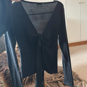 Svart långärmad topp med knyt - Svart långärmad topp från Ginatricot med djup v-ringning och knyt framtill. Tunn och stretchig, perfekt att styla med jeans eller kjol. Använd ett fåtal gånger men är i nyskick!