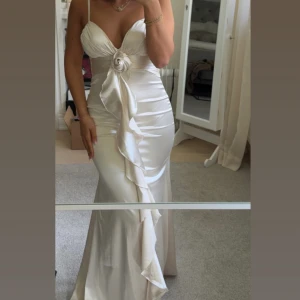 Vit långklänning i satin från Bella Barnett - Elegant vit långklänning i satin från Bella Barnett med smala axelband, djup v-ringning och dekorativ volang framtill. Klänningen har en stor rosettdetalj vid bysten och figurnära passform som framhäver siluetten.