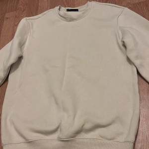 Beige sweatshirt med rund hals - En stilren beige sweatshirt med rund hals och långa ärmar. Tröjan har ribbade muddar vid ärmslut och nederkant, vilket ger en klassisk och enkel look. Perfekt att matcha med jeans eller mjukisbyxor.