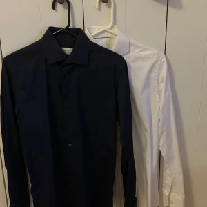 Mörkblå och vit skjorta från Eton - Två stilrena skjortor från Eton i storlek 38, en Mörkblå och en vit. Båda har klassisk krage, lång ärm och knäppning framtill. Slim fit-modell som sitter snyggt och passar till många tillfällen. Perfekt för dig som gillar en clean och tidlös look.