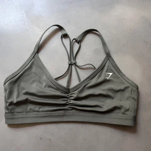 Olivgrön sport-bh från Gymshark - Säljer en olivgrön sport-bh från Gymshark med smala axelband och snygg knytdetalj i ryggen. Bh:n har lätt rynkning framtill och är tillverkad i ett mjukt, stretchigt material som passar perfekt för träning. Kan köpas med shortsen för 350kr 💕