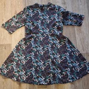 Blommig klänning från Zara Basic - Säljer en blommig klänning från Zara Basic med trekvartsärm och rund halsringning. Klänningen har en utställd kjol och ett färgglatt blommönster i grönt, rött, blått och vitt. Perfekt för dig som gillar färg och mönster!