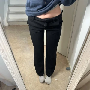 Svarta kostymbyxor med låg midja - Säljer ett par svarta kostymbyxor med låg midja och raka ben. Byxorna har bälteshällor och knäppning fram. Perfekta för en stilren och klassisk look.