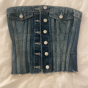 Jeanskorsett från ZARA med knappar - Säljer en snygg jeanskorsett från ZARA i tvättad blå denim. Toppen har silverfärgade knappar framtill, två dekorativa fickor och rå kant nertill. Perfekt för att skapa en cool och trendig look.