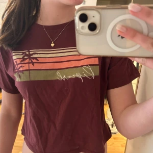 Vinröd croppad t-shirt från Rip Curl - Säljer en vinröd croppad t-shirt från Rip Curl med tryck av palmer och färgade ränder i orange, rosa och olivgrönt på bröstet. T-shirten har korta ärmar och är tillverkad i mjuk bomull. Perfekt för sommaren!