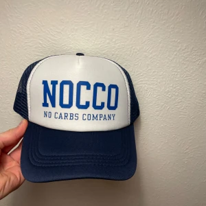 Blå och vit keps från NOCCO - Snygg blå och vit keps från NOCCO med tydlig logga framtill. Kepsen har nät på sidorna och baksidan för extra ventilation och en böjd skärm. Perfekt accessoar för en sportig look. Limiterad till få antal och aldrig använd 