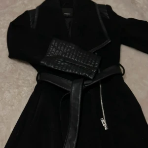 Svart kappa med skinn-detaljer och bälte - Säljer en svart kappa med snygga skinn-detaljer på axlar och ärmslut. Jackan har ett bälte i midjan, Perfekt för våren men också hösten.