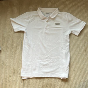 Vit piké t-shirt från Levi's - Säljer en klassisk vit piké t-shirt från Levi's med krage och knappar framtill. T-shirten har korta ärmar och en broderad Levi's-logga på bröstet. Storlek 164