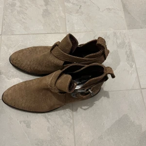 Bruna boots med ormskinnsmönster och spänne - Säljer ett par bruna boots med ormskinnsmönster och dekorativt spänne vid ankeln. Skorna har spetsig tå och är tillverkade i ett material som liknar mocka. Perfekta för dig som vill sticka ut med en unik detalj.