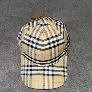 Beige rutig keps från Burberry - Säljer en klassisk keps från Burberry i beige med svart, vit och röd rutig design. Kepsen har böjd skärm och justerbar rem baktill. Perfekt accessoar för en stilren look.