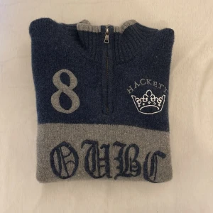 Blå halvzip tröja från Hackett London - Säljer en blå och grå halvzip tröja från Hackett London i 100% merinoull. Tröjan har hög krage, dragkedja och broderade detaljer med siffran 8, OUBC och en krona. Union Jack-flagga på ärmen. Perfekt för dig som gillar klassisk brittisk stil.