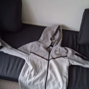 Nike tech fleece - Säljer en ljusgrå hoodie från Nike med hel dragkedja och huva. Jackan har svarta detaljer längs dragkedjan och fickor på sidorna. Perfekt för en sportig och avslappnad stil. Priset är ej hugget i sten. STORLEK S