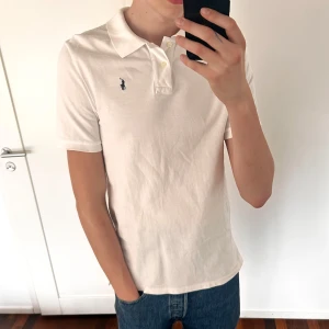 Vit pikétröja från Polo Ralph Lauren - (OBS, Piken är storlek XL för barn så motsvarar S-M för vuxen) Klassisk vit pikétröja från Polo Ralph Lauren med korta ärmar och broderad logga på bröstet. Tröjan har krage och knappar framtill, perfekt för en clean och stilren look.