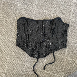 Svart paljettkorsett med snörning - Säljer en svart korsett topp täckt med glittriga paljetter. Toppen har snörning i ryggen för justerbar passform och är croppad i modellen. Perfekt för dig som vill sticka ut med en trendig och festlig look🌸