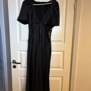 Balklänning  - Säljer en svart långklänning från Boohoo med djup v-ringning och spetsdetaljer i sidorna. Klänningen har korta ärmar och en elegant, figurnära passform. Perfekt för dig som vill sticka ut med en stilren och sofistikerad look.