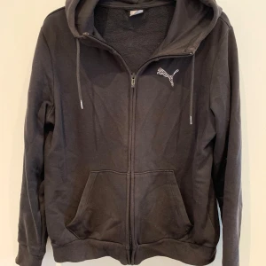 Svart hoodiejacka från Puma - Svart hoodiejacka från Puma med dragkedja framtill och vit broderad logga på bröstet. Jackan har huva med dragsko och två fickor framtill. Perfekt för en avslappnad stil.