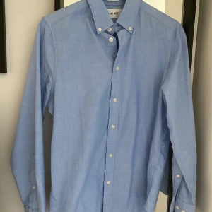 Skjorta - Säljer en klassisk ljusblå skjorta från East West i ekologisk bomull. Skjortan har button-down krage, långa ärmar och vita knappar framtill. Perfekt för en stilren och enkel look.