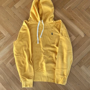 Gul hoodie från Polo Ralph Lauren - Säljer en gul hoodie från Polo Ralph Lauren med klassisk broderad logga på bröstet. Tröjan har huva med vita snören och en stor magficka. Perfekt för dig som gillar stilrena och bekväma plagg.