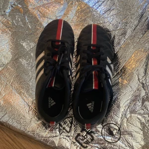 Adidas fotbollsskor med röd och vit detalj - Svarta Adidas fotbollsskor med vita ränder på sidorna och röda samt vita detaljer längs tungan och hälen. Klassisk design med snörning och rund tå, perfekt för dig som gillar sportig stil.