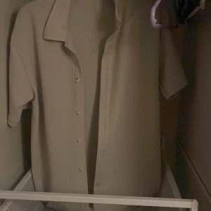 Beige skjorta med kort ärm och knappar - Säljer en stilren beige skjorta med korta ärmar och knäppning framtill. Skjortan har en klassisk krage och är tillverkad i ett mjukt material med diskret struktur. Perfekt för en avslappnad och trendig look. Den här skjortan är i ett mycket bra skick och hade passat Perfekt nu till sommaren. Kan skicka fler bilder privat 