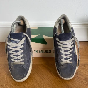 Blå sneakers från The Gallerist - Säljer ett par blå sneakers från The Gallerist med vita detaljer och snörning. Skorna har rund tå och platt sula. Ovandelen är i mocka och de har en vit hälkappa med logotyp. Perfekta för en avslappnad stil.