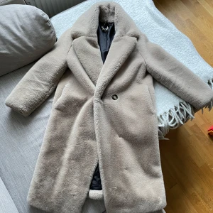 Beige pälsjacka med bred krage - Säljer en lång, beige pälsjacka med bred krage och en knapp framtill. Jackan har långa ärmar och är helfodrad med svart innerfoder. Perfekt för kyliga dagar när du vill vara både varm och snygg.