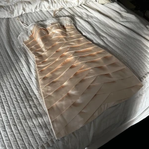 Champagnefärgad miniklänning med fransdetalj - Säljer en champagnefärgad miniklänning med tunna axelband och fransar upptill. Klänningen har omlottliknande banddetaljer i satin och justerbara axelband. Perfekt för dig som vill sticka ut med en elegant och trendig look.