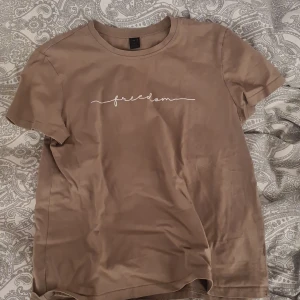 Beige t-shirt med trycket 'freedom' från SHEIN - Beige t-shirt från SHEIN med vit text 'freedom' på bröstet. Klassisk rund halsringning och korta ärmar. Tillverkad i mjuk bomull, perfekt för en avslappnad stil.