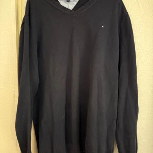 Mörkblå v-ringad tröja från Tommy Hilfiger - Klassisk mörkblå v-ringad tröja från Tommy Hilfiger med diskret logga på bröstet. Tröjan har långa ärmar och ribbade muddar vid hals, ärmslut och nederkant. Perfekt för en stilren och enkel look. Det är storlek 176 i barnstorlek men den passar även S