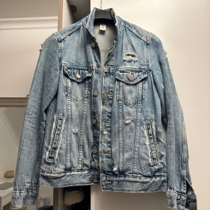 Blå jeansjacka från &Denim - Säljer en klassisk blå jeansjacka från &Denim i storlek XS. Jackan har knappar framtill, bröstfickor med lock och slitna detaljer för en cool look. Perfekt att slänga över en hoodie eller t-shirt.