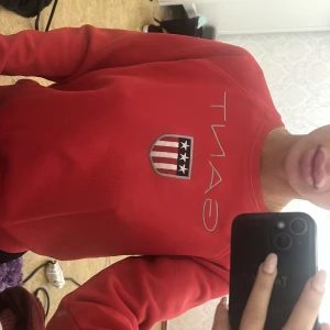 Röd sweatshirt från Gant - Röd sweatshirt från Gant med stort tryckt logotyp och sköld på bröstet. Tröjan har rund halsringning och långa ärmar. 