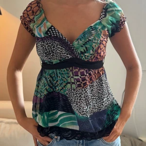 Färgglad omlottopp med djurmönster - Säljer en färgglad omlottopp med blandade mönster i grönt, svart, lila och beige. Toppen har korta ärmar, v-ringning och svarta spetsdetaljer nertill. Mönstret är en mix av leopard, rutigt och grafiska former. Perfekt för dig som gillar unika plagg!