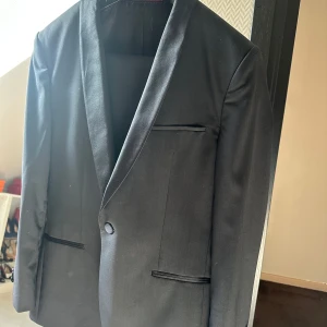 Svart smoking inkl byxor från Hugo Boss - Säljer en stilren svart smoking från Hugo Boss med klassisk passform och smala slag. Kavajen har tre fickor framtill och en innerficka, samt elegant foder med röda detaljer. Perfekt för bal eller fest. Endast använd en gång till bal. Pris går att diskuteras.