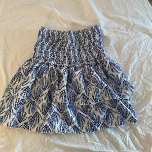 Mini kjol - Supersöt kortkjol i vitt med blått bladliknande mönster och bred smock i midjan. Kjolen har volang och är fodrad för extra komfort. Perfekt för en fräsch och somrig look! Midjebandet kan man göra mindre och det är även stretchigt. Det finns inbyggda shorts. Hör av er via frågor och mer bilder💕