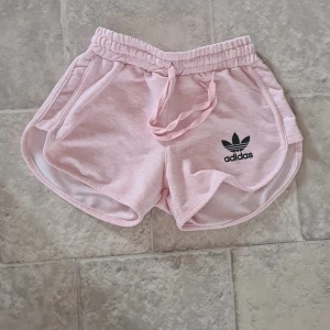 Ljusrosa shorts från Adidas - Säljer ett par ljusrosa shorts från Adidas med svart logga på ena benet. Shortsen har elastisk midja med snörning och är tillverkade i mjukt bomullsmaterial. Perfekta för träning eller varma sommardagar.