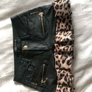 Svart minikjol med leopardvolang - Cool svart minikjol från Gogo Jeans med dragkedjedetaljer och volang i leopardmönster längst ner. Kjolen har fickor både fram och bak samt guldiga knappar och dragkedjor. Perfekt statement piece för dig som vill sticka ut.