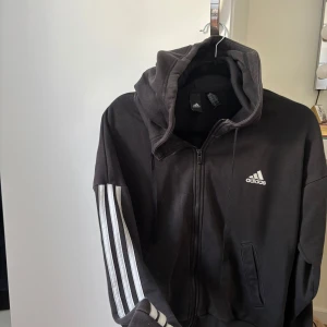 Svart Adidas hoodie med dragkedja - Svart hoodie från Adidas i storlek M med klassiska vita ränder längs ärmarna och Adidas-logga på bröstet. Tröjan har huva med snörning, dragkedja framtill och två fickor. Perfekt för en sportig och avslappnad stil.