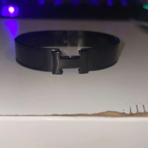 Svart H-armband i metal - Stilrent svart armband med ikoniskt H-spänne framtill. Armbandet är tillverkat i metall och har en enkel, modern design som passar perfekt till din outfit. Lätt att ta av och på tack vare det smidiga spännet.