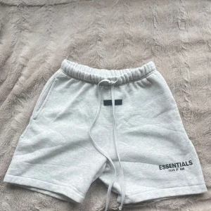 Grå shorts Essentials Fear of God XS - Ljusgrå mjuka shorts från Essentials Fear of God med elastisk midja och snörning. Klassisk logga tryckt på vänster ben, två sidofickor och relaxed fit. Perfekta för chill dagar eller när du vill ha en clean streetwear-look.