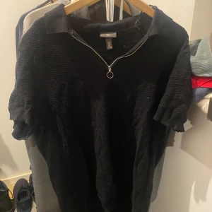 Svart pikétröja med half zip från H&M - Svart pikétröja från H&M i slim fit med korta ärmar och snyggt vågigt strukturmönster. Tröjan har en klassisk krage och en half zip-dragkedja med ringdetalj framtill. Perfekt för dig som gillar stilrena och moderna plagg.