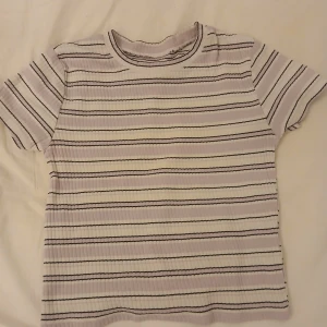 Randig ribbad topp från H&M - Superfin ribbad topp från H&M i ekologisk bomull. Den har lila, vita och svarta horisontella ränder och en klassisk rund hals. Kortärmad och mjuk, perfekt för en chill och stilren look.