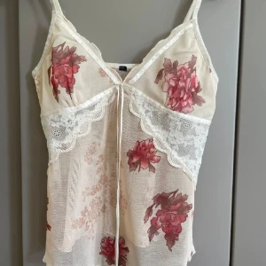 Blommig spetslinnetopp med rosett - Romantisk linne-topp i ljusbeige mesh med stora rosa och röda blommor. Spetsdetaljer och liten rosett framtill, smala axelband och v-ringning. Perfekt för lager-på-lager eller som statement-piece till jeans.